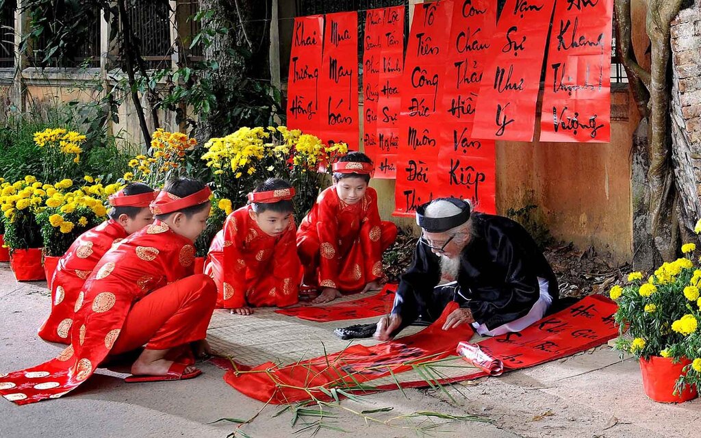 A calligrapher in traditional dress writes red Tet banners as children in red ao dai watch