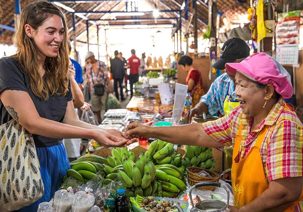 A traveller pays a smiling vendor at a banana and vegetable stall in a local market