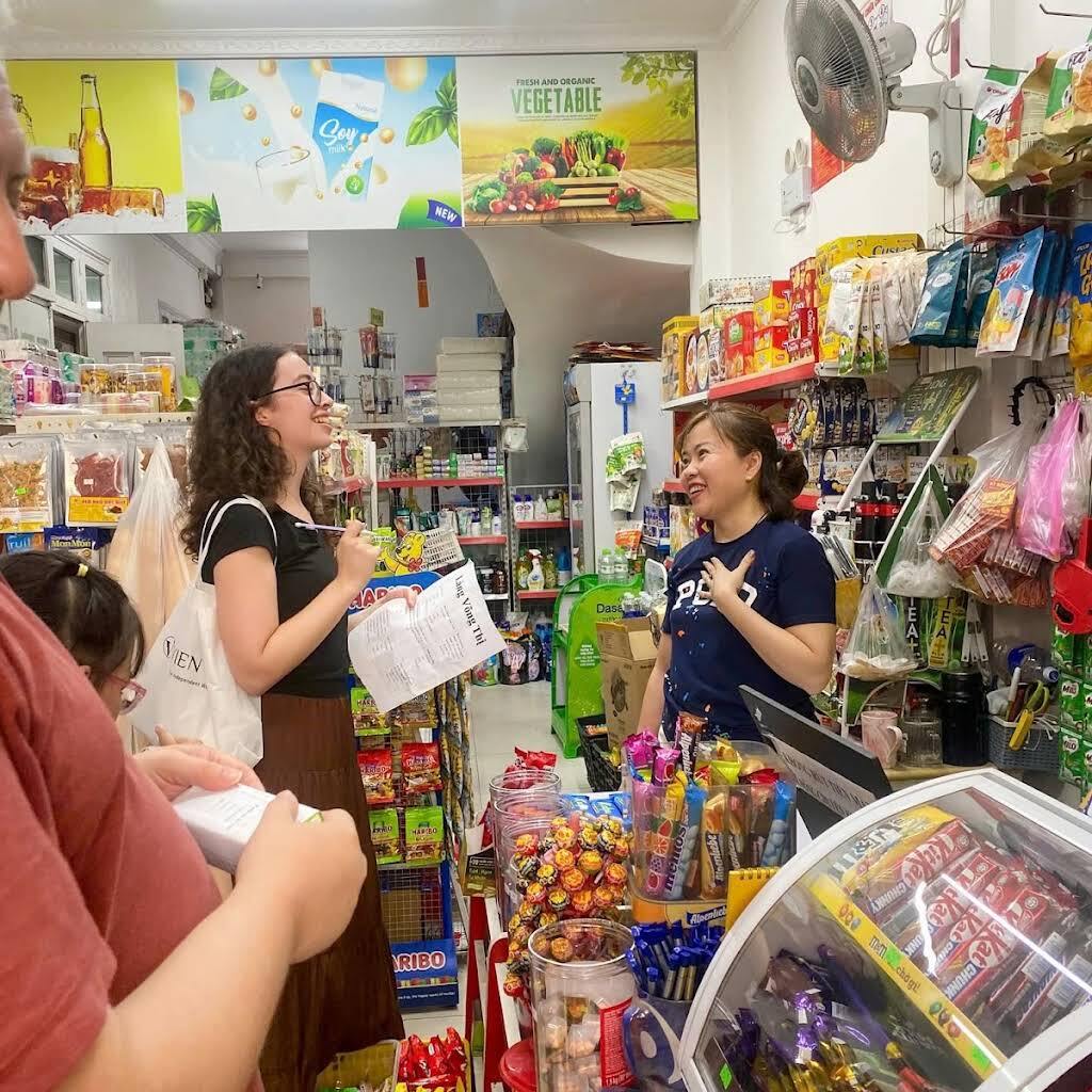 A visitor with a shopping list asks a shop assistant about prices in a small grocery store